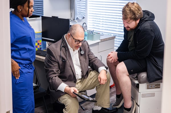 Dr. Wael Aboughali performs a metabolic examination while discussing symptoms with a patient during a comprehensive primary care appointment, with a medical assistant present.